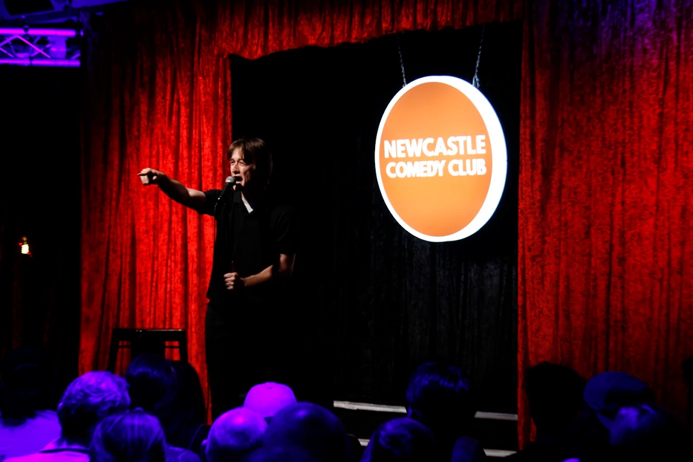 Comedian performing on stage at Newcastle Comedy Club with red curtains and club logo